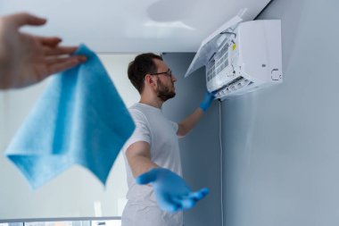 Görüntü, bir klima ünitesine hizmet eden eldivenli birini gösteriyor. Mavi bir temizleme bezini uzatan başka bir kişi daha görülüyor, bu da bakım için işbirliği yaptığını gösteriyor..