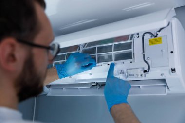 Görüntü, klima sistemi üzerinde bakım yapan, filtreleri ayarlayan ve servis sırasında güvenlik ve temizlik için koruyucu eldivenler kullanan bir teknisyeni resmediyor..