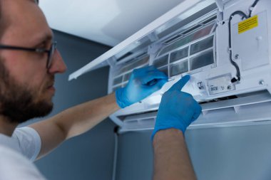 Mavi eldivenli bir teknisyen klima ünitesini temizliyor. İçerideki filtreyi temizliyor. Modern yaşam ortamında hava kalitesini ve verimliliğini korumaya çalışıyor..