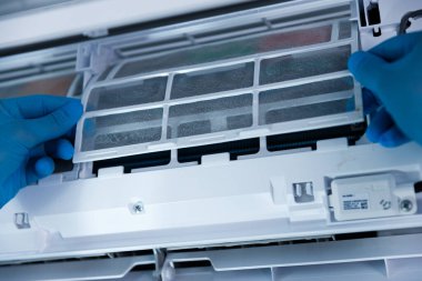 Yakın plan bir görüntüde mavi eldiven giyen bir kişi, sağlık ve işlevsellik için düzenli temizliğin önemini vurgulayarak, bir klima filtresini ustalıkla kaldırıyor..