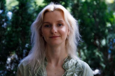 Görüntü, açık sarı saçlı bir kadının yakın plan portresini gösteriyor. Arkaplanda bulanık yeşil bir yeşillik var. Huzurlu ve huzurlu bir ambiyansı çağrıştırıyor..