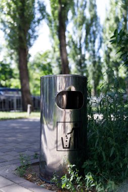 Bu resim, halka açık alanlarda daha temiz bir ortamın atık imhasını teşvik eden bir tabela ile canlı yeşilliklerin arasına yerleştirilmiş parlak paslanmaz çelik bir çöp tenekesini gösteriyor..