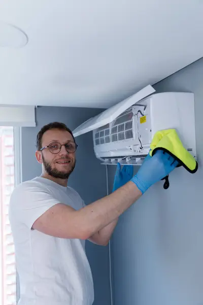 Beyaz tişörtlü ve mavi eldivenli gülümseyen bir adam havalandırma ünitesini temizliyor. Dikkati, rahat bir yaşam ortamını korumaya olan bağlılığını gösteriyor..