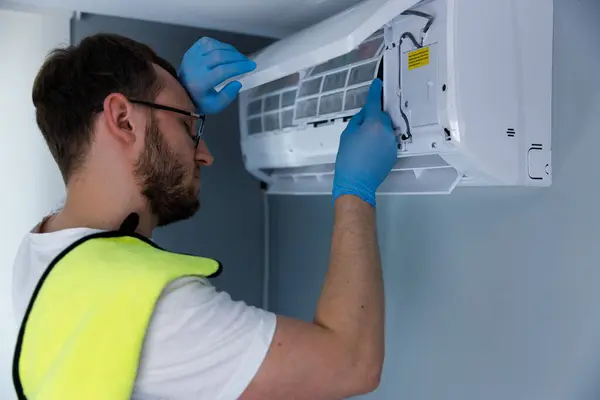 Görüntü, klima sistemini tamir etmeye odaklanmış bir teknisyeni yakalıyor. Mavi eldiven ve parlak sarı bir yelek giyiyor. Birim bileşenleri üzerinde çalışırken güvenliği vurguluyor..