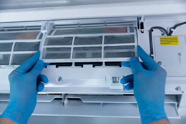 Görüntüde, bir klima ünitesinin içinden bir filtre çıkarırken, verimli soğutma için uygun bakım ve bakım prosedürlerini vurgulayan mavi eldivenli bir çift el görülüyor..