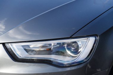 Bu görüntü, bir arabanın ön alanının yakın görüntüsünü sunuyor, şık bir far tasarımı, pürüzsüz yüzey ve etrafındaki gökyüzü ışığı tarafından tamamlanmış ayrıntılı yansımaları gösteriyor..