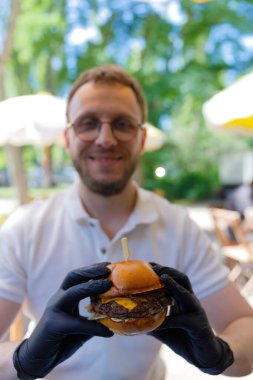 Eldiven takan neşeli bir birey, canlı bir açık hava ortamının ortasında ağız sulandıran bir çizburger tutuyor. Mutfak zevkini tatmak ve yemeği keyifli kılmak için heyecanlarını sergiliyor..