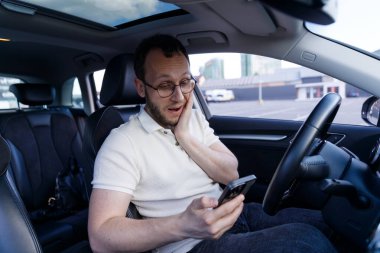 Arabada şaşırmış bir adam akıllı telefonunu kontrol ediyor, şok olmuş bir ifadeyle karışık duygular gösteriyor, araba camlarından şehir manzarası çerçevesi ile çerçevelenmiş..
