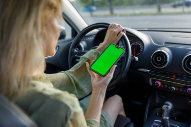 Bu resimde, bir kadın bir elinde yeşil ekranlı akıllı bir telefonu tutarken araba kullanmaya odaklanmış, teknoloji ve güvenli sürüş uygulamaları arasındaki dengeyi vurguluyor..