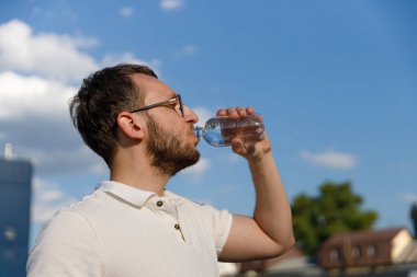 Görüntü, açık mavi gökyüzü ve güzel bulutlarla çevrili sıcak havanın tadını çıkarırken plastik şişeden su içen bir adamı yakalıyor..