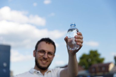 Görüntüde neşeli bir genç adam berrak suyla dolu plastik bir şişe tutuyor, canlı bir gökyüzünün zeminine kurulmuş, bulutlar sahneye cazibe katıyor..