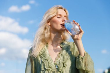 Berrak mavi gökyüzü ve yumuşak bulutlarla çevrili temiz bir şişeden bir yudum su içen bir kadının canlandırma ve canlılığın özünü vurguladığı sakin bir sahne..