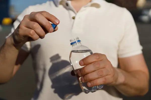 Kapağı açarken elinde temiz bir plastik su şişesi tutan kişinin yakın plan görüntüsü. Güneş, açık hava ortamında yere gölge düşürüyor..