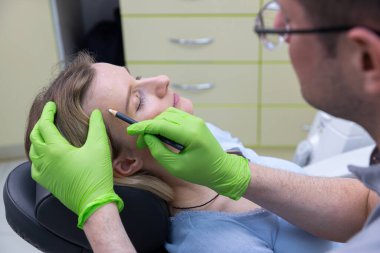 Bu resimde, eğitimli bir profesyonel, parlak ve temiz bir klinik ortamda kusursuz bir taslak çizmek için kozmetik kalem kullanan bir hastanın görünümünü geliştirmeye hazırlanıyor..