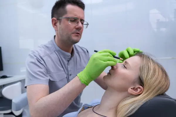 Eldivenlerde yetenekli bir kozmetik uzmanı, hastaların iyi ışıklandırılmış ve modern bir klinik ortamında estetik bir prosedür için yüzlerini işaretlemek için bir kalem kullanır..