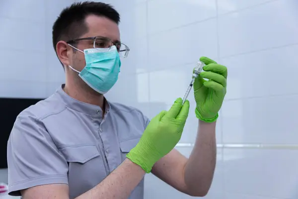 Gri üniformalı ve yeşil eldivenli bir sağlık çalışanı bir aşı hazırlar, yüz maskesi takarken güvenliği sağlar, sağlık ve hijyenin tıbbi ortamlardaki önemini vurgular.