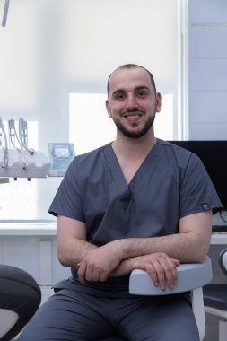 Önlüklü bir dişçi diş muayenehanesinde rahatça oturuyor ve dostane bir tavır sergiliyor. Klinik, modern diş aletleri ve parlak ışıklandırmalarla donatılmıştır..