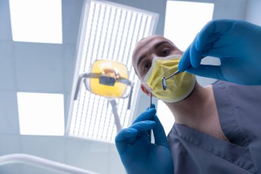 Gri üniformalı ve koruyucu donanımlı bir dişçinin iyi aydınlatılmış bir klinikte diş aletlerini tutarak oral sağlık ve profesyonel bakımın önemini vurguladığı yakın plan bir perspektif..