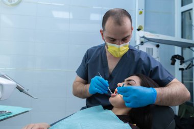 Cerrahi maskeli ve eldivenli bir diş hekiminin parlak, hijyen kliniğinde ihtiyacı olan hastalara dikkatlice bakması, ağız sağlığı hizmetlerindeki güvenlik ve kalite hizmetlerine odaklandığını göstermektedir..