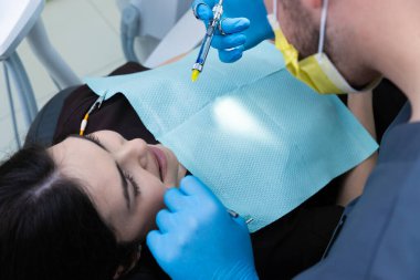 Bu görüntü, dişçi olarak dişçi koltuğunda rahat bir şekilde yaslanan genç bir kadını, iyi aydınlatılmış modern bir klinik ortamında hassas araç ve teknikler kullanarak yakalar..