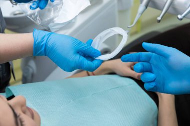 Bu diş ortamında, mavi eldivenli bir diş hekimi bir diş aletini hazırlarken, diğer bir kişi, hasta, muayene için bir tedavi koltuğunda yatmaktadır..