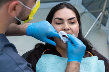 Mavi eldivenli bir diş hekimi, diş sağlığı kliniğinde hastaya yardım ederken güvenlik protokollerine bağlı kalır ve diş bakımında hijyenin önemini vurgular..