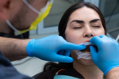 Koruyucu giysili bir dişçi bir hastaya sözlü muayene yapıyor. Prosedür, diş aletlerinin dikkatli bir şekilde kullanılmasını ve klinik ortamda hasta konforunun sağlanmasını içeriyor..