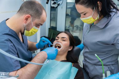 Bir diş hekimi ve dişçi asistanı, sağlık kontrolünde bir hasta üzerinde birlikte çalışırlar, takım çalışması ve güvenlik protokollerine uyulduğunu klinik bir ortamda maskeler ve eldivenlerle gösterirler..