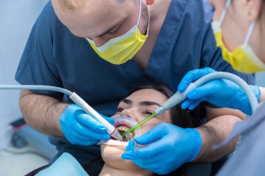Bu diş sağlığı kliniği sahnesinde, iki uzman bir hasta üzerinde dikkatle bir prosedür yürütüyor, oral sağlık hizmetleri için özel araçların ve güvenlik önlemlerinin kullanımını gösteriyorlar..