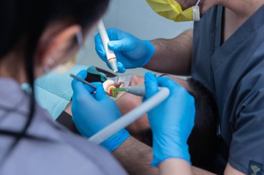 Bu görüntüde, bir hastanın iki diş hekiminden bakım gördüğü, modern araçların kullanımını ve diş hekimliğinde takım çalışmasının önemini gösteren bir diş prosedürü yer alıyor..