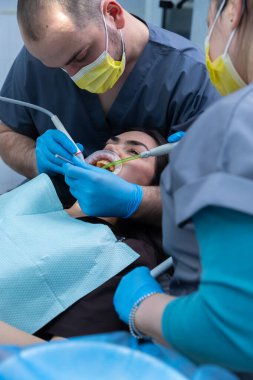 Bir diş kliniğinde, koruyucu giysiler ve maskeler giyen iki diş hekimi tedavi gören bir hastaya, takım çalışmasına ve diş sağlığı hizmetlerinin önemine odaklanır..