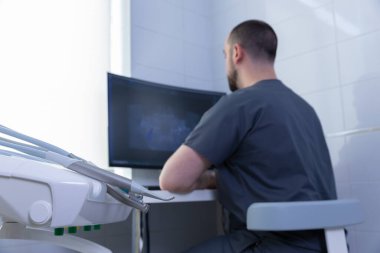 Önlük giymiş bir diş hekimi, parlak bir diş muayenehanesinde bulunan bir bilgisayar monitöründeki dijital röntgen görüntülerini dikkatle inceliyor..