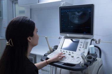 Bu resimde, uzun siyah saçlı ve saç aksesuarlı bir kadın, temiz bir tıbbi tesiste teşhis imgelerini gösteren bir ultrason makinesini çalıştırmaya odaklanıyor..