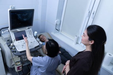 Bir tıp uzmanı klinik ortamda bir hasta üzerinde ultrason muayenesi gerçekleştirirken, diğer bir personel de sağlık hizmetlerinin kalitesini vurgulayarak bilgiyi gözlemler ve iletir..