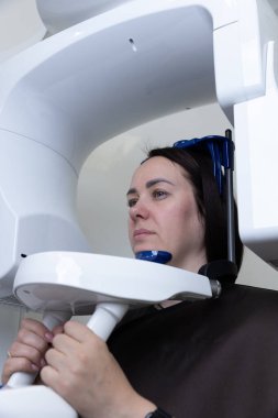 Bu görüntüde diş röntgeninden geçen bir hasta görülüyor. Oturmuş, tıbbi teşhisler için detaylı görüntüler yakalayan beyaz bir makineye tutunuyor..