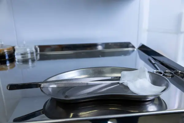 Görüntü, temiz paslanmaz çelik cerrahi tepsiyi cımbız ve gazlı bez de dahil olmak üzere hassas aletlerle gösteriyor. Hijyen üzerine odaklanmış titiz bir tıbbi çalışma alanını yansıtıyor..