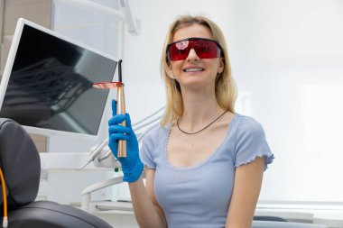 Görüntü, diş kliniğinde genç bir kadını gösteriyor. Elinde diş aletini tutarken özgüvenle gülümsüyor. Koruyucu gözlük takıyor, hasta bakımında profesyonelliği simgeliyor..