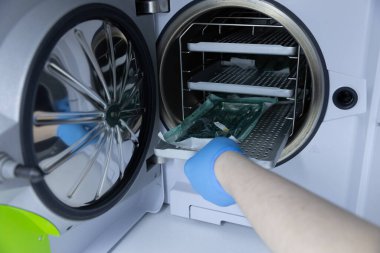 Görüntü, sterilizatörden sterilize edilmiş ürünler alan, sağlık hizmetlerinde hijyen standartlarını korumak için hassasiyet ve bakım gösteren mavi eldivenler giyen bir kişiyi yakalıyor..