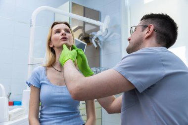 Bir diş kliniğinde dişçi, genç bir kadının çene hattını ve çenesini eldivenli ellerle inceler. Kişisel bakım ve profesyonel diş sağlığı uygulamalarının önemini gözler önüne serer..