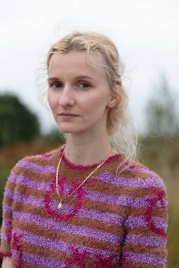 Görüntü, canlı çizgili bir kazak ve altın zincirli kolye giymiş, bulutlu bir günde yeşil bir manzaranın ortasında sakin bir aura yayan yumuşak hatlara sahip genç bir kadını yakalıyor..