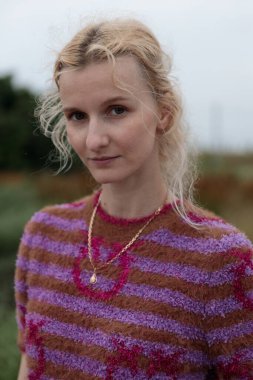 Bu çarpıcı portre kıvırcık, sarı saçlı, parlak çizgili kazak giymiş genç bir kadını gösteriyor. Arka plan hafifçe bulanık, karamsar bir atmosfer yaratıyor..