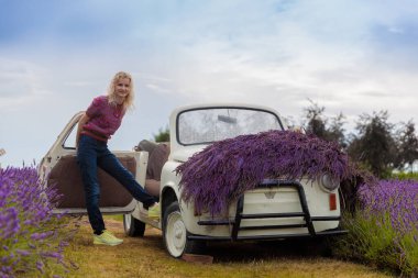 Çekici genç bir kadın süslü, lavantalı bir arabadan neşeyle iniyor. Araba, canlı bir lavanta tarlasına karşı kurulmuş. Resimli bir doğal manzara yaratıyor..