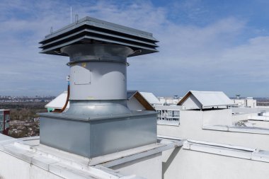 Görüntü, bina için hava akımını optimize etmek üzere tasarlanmış büyük bir vantilatör deliğinin yer aldığı bir çatı havalandırma sistemi sergiliyor. Aşağıda, kentsel çatılar uzak bir ufuk çizgisine doğru uzanır ve manzarayı yükseltir..