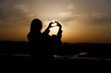 Göz kamaştırıcı bir günbatımı sahnesi, bir bireyin siluetini gösteriyor. Elleriyle bir kalp şekli yaratıyor. Gökyüzündeki sıcak turuncu ve sarı renklerle güzel bir tezat oluşturuyor..