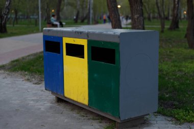 Bir sıra üç farklı geri dönüşüm kutusu, renkli mavi, sarı ve yeşil, park yolu boyunca yerleştirilmiş, ziyaretçileri doğanın tadını çıkarırken atıklarını sorumlu ve sürdürülebilir bir şekilde ortadan kaldırmaya teşvik ediyor..