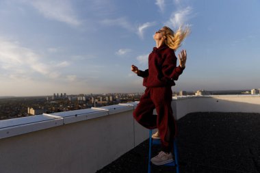 Görüntü, koyu kırmızı bir elbise giyen neşeli bir genç kadını yakalıyor, çatıda mavi bir sandalyede duruyor, serinletici rüzgarda şehrin arka planına doğru güneşleniyor..