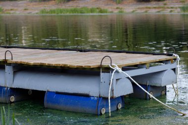 Tahtadan yapılmış ve varillerle desteklenen yüzen bir iskele dingin suyun üzerinde duruyor. Sakin ortam barışçıl yansımalar ve yemyeşil bitki örtüsüyle karakterize edilir..