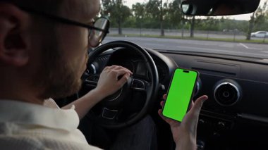 Bir kişi arabanın içinde canlı yeşil bir ekranın bulunduğu akıllı telefonu aktif olarak kullanırken görülüyor, araba kullanırken teknolojiyle bağlantısını gösteriyor..