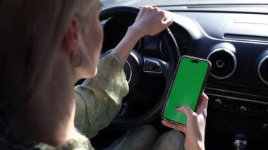 Bir kişi elinde akıllı bir telefonla yeşil ekran gösteren bir arabayı tehlikeli bir şekilde kullanıyor, dikkat dağıtma ve direksiyon başında birden fazla iş yapma sorunlarını vurguluyor..