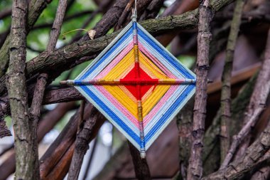 Renkli bir geometrik tekstil asılı, birbirine dolanmış ağaç dallarının bir zeminine karşı sergileniyor, sanatsal tasarımı ve canlı renkleri vurgulanıyor, doğa ve zanaatkarlığı güzelce harmanlıyor..